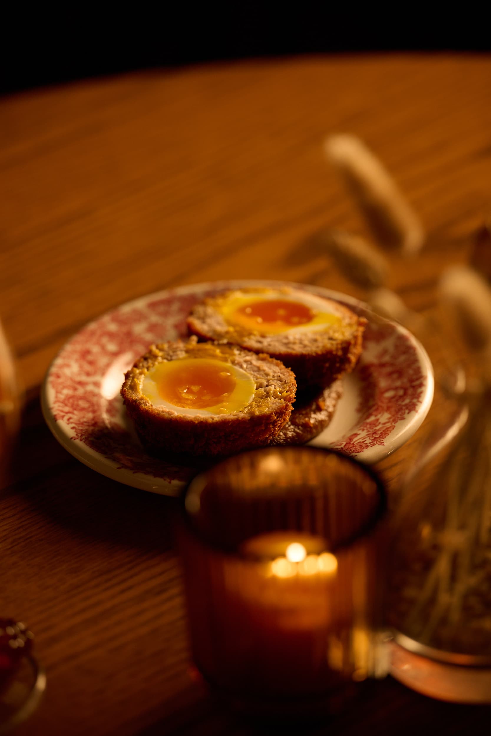 A MacTaverne scotch egg cut in half, runny yolk, on a red floral-pattern plate, by candlelight