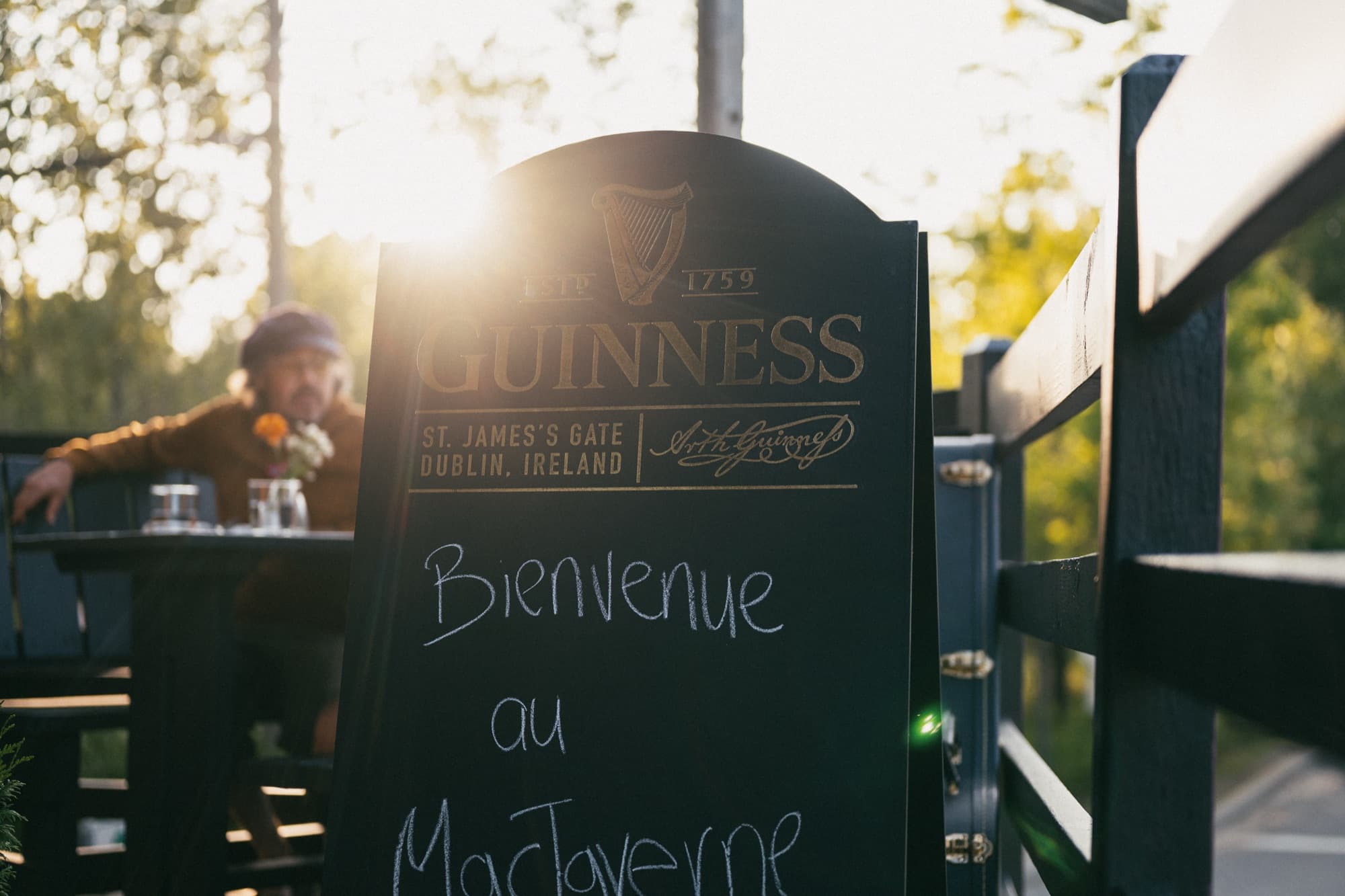 A Guinness chalkboard reading “Bienvenue au MacTaverne” on the terrasse at sunset, a guest seated at a table behind it